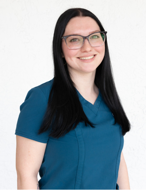 Woman wearing glasses and blue scrubs smiling at camera in professional portrait setting.