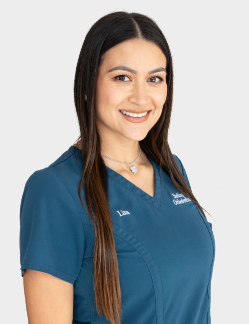 Female dental professional in blue scrubs smiling at camera against neutral background.