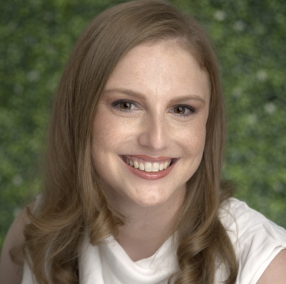 Young woman smiling at camera wearing white shirt against green foliage background.